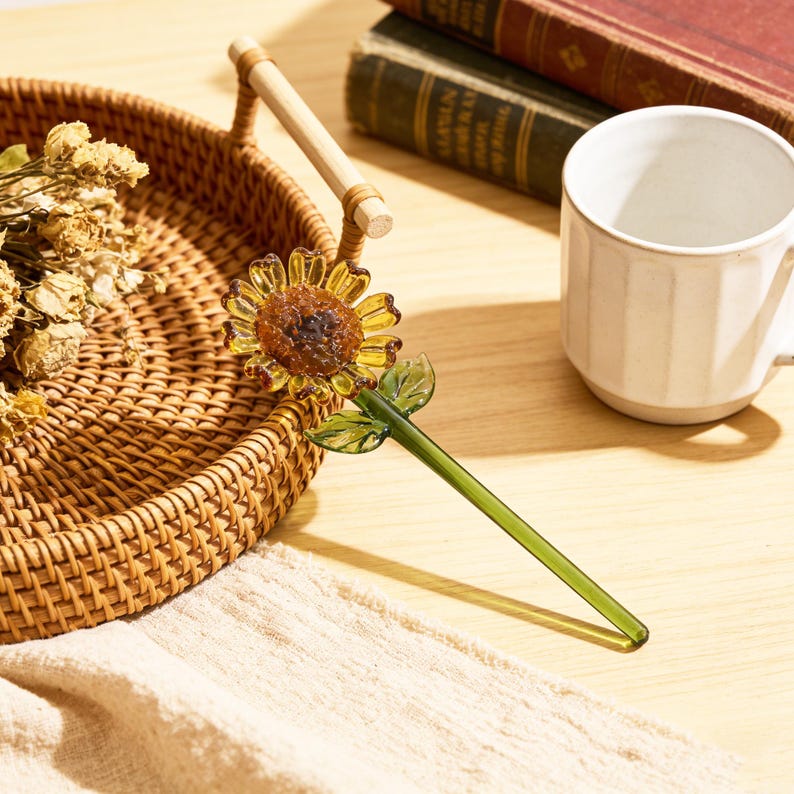May include: A decorative glass sunflower with a yellow and brown flower head, green stem, and leaves. The sunflower is next to a woven basket, a white mug, and books. The sunflower is a unique home decor item.
