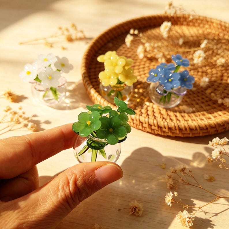 May include: Miniature glass flower bouquets in clear vases. The bouquets feature white, yellow, blue, and green flowers. The vases are arranged on a woven tray, with one held in a hand. The flowers are detailed and delicate.