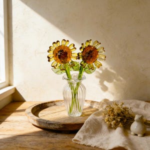 May include: Two glass sunflower stems in a clear, ribbed vase. The sunflowers have yellow petals and brown centers. The vase sits on a wooden tray with a white cloth and small ceramic figurine.
