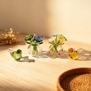 May include: Glass bird figurines and miniature vases with colorful flowers. The vases are clear with yellow and orange polka dots. The birds are green and yellow. The flowers are blue, purple, and yellow. The items are on a wooden surface.