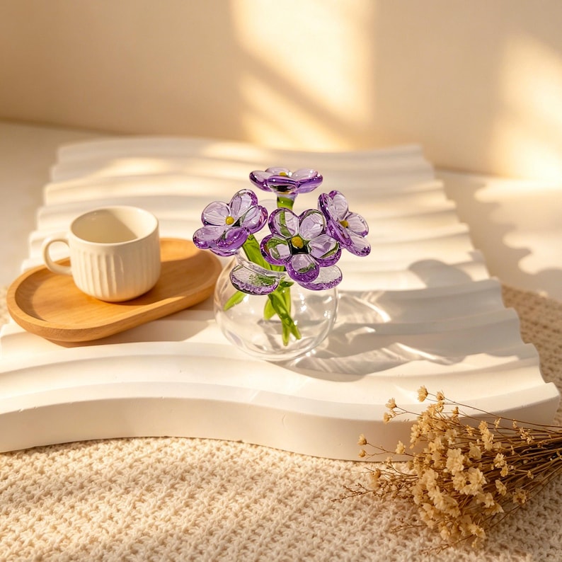 May include: A small, clear glass vase holds a cluster of purple glass flowers with green stems and yellow centers. The vase sits on a white, wavy tray, next to a small white teacup and a wooden saucer. The scene is bathed in soft light.