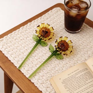 May include: Two glass sunflower-shaped drink stirrers with yellow petals and green stems and leaves. The stirrers are on a white woven placemat, next to a glass of iced coffee and an open book.