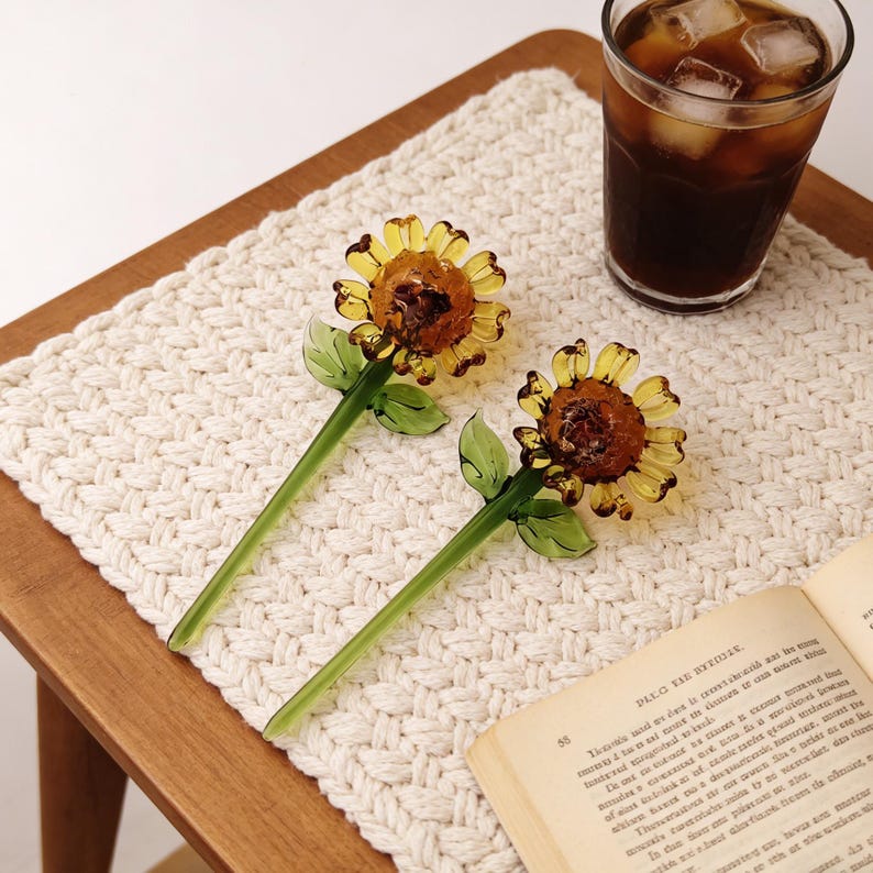 May include: Two glass sunflower-shaped drink stirrers with yellow petals and green stems and leaves. The stirrers are on a white woven placemat, next to a glass of iced coffee and an open book.