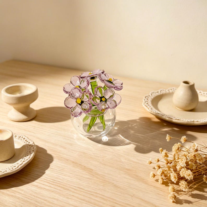 May include: A small glass vase holds a bouquet of delicate, light purple flowers with yellow centers and green stems. The vase sits on a light wooden surface, with other decorative items nearby, including a small, off-white vase and a candle holder.