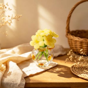 May include: A small, clear glass vase with yellow flower-shaped decorations and stems, holding several yellow flower blooms. The vase sits on a wooden surface, with a woven basket and a cream-colored cloth in the background.