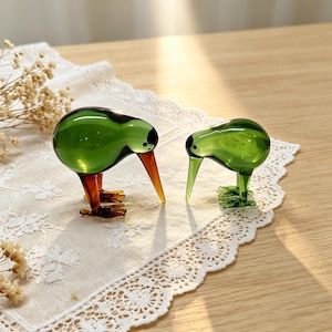 May include: Two glass kiwi bird figurines. The larger figurine features a translucent green body with dark brown legs and a long, pointed brown beak. The smaller figurine is entirely translucent green with a long, slender beak and small feet.