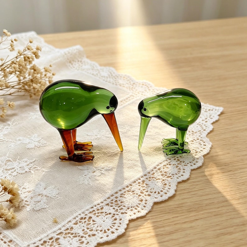 May include: Two glass kiwi bird figurines. The larger figurine features a translucent green body with dark brown legs and a long, pointed brown beak. The smaller figurine is entirely translucent green with a long, slender beak and small feet.