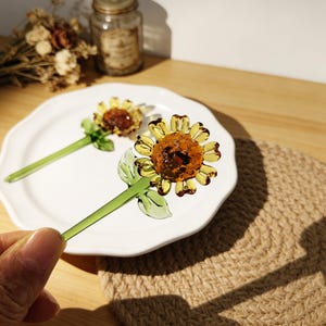 May include: Two glass sunflower ornaments with yellow petals and brown centers rest on a white plate. The stems and leaves are green. The sunflowers are decorative and add a touch of nature to any space.