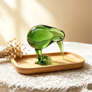May include: A translucent green glass kiwi bird figurine with a long, pointed beak and detailed feet, standing on a smooth, rectangular light-colored wooden tray. The glass bird has a rounded body and a small, dark, circular eye detail.