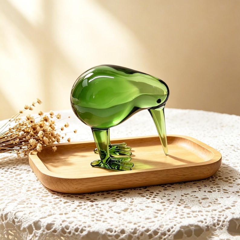 May include: A translucent green glass kiwi bird figurine with a long, pointed beak and detailed feet, standing on a smooth, rectangular light-colored wooden tray. The glass bird has a rounded body and a small, dark, circular eye detail.