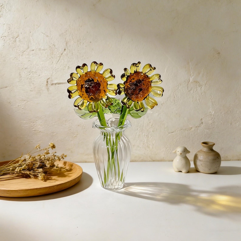 May include: Two glass sunflowers with yellow petals and brown centers are displayed in a clear, fluted glass vase. The stems are green. The vase sits on a white surface with a wooden plate and small ceramic figurines in the background.
