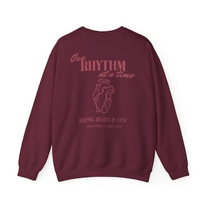 May include: A maroon long-sleeve sweatshirt featuring a pink anatomical heart graphic on the back. The text reads One RHYTHM at a time, KEEPING HEARTS IN SYNC, and MIDRA APPAREL CO. ESTD 2024. The design is centered on the back of the garment.