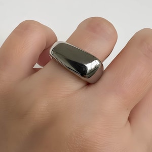May include: A polished, silver-toned ring with a rectangular, rounded top. The ring is smooth and reflective, catching the light. The jewelry piece is worn on a finger.