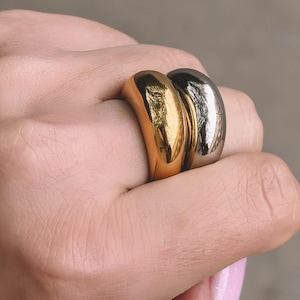 May include: Two chunky rings, one gold and one silver, are displayed on a hand. The rings have a smooth, rounded design. The gold ring is on the index finger, and the silver ring is on the middle finger.