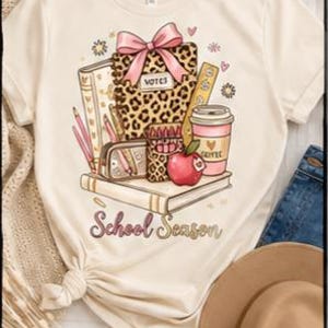 May include: A cream-colored t-shirt featuring a graphic print of school supplies including a leopard print notebook with a pink bow, a red apple, a coffee cup, pencils, and books. The text "School Season" is printed in a cursive font below.