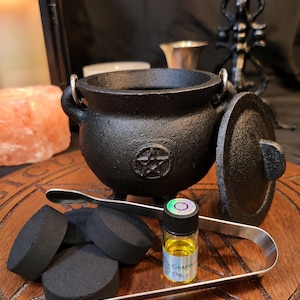 May include: A black cast iron cauldron with a pentagram symbol, a matching lid, and a metal handle. Included are four black charcoal discs, a pair of silver-toned metal tongs, and a small glass bottle containing yellow Grapes Oil.