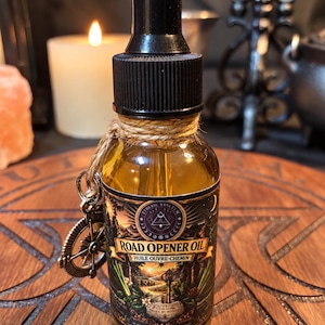 May include: A clear glass dropper bottle filled with golden oil, featuring a black cap and a twine-wrapped neck with a small metal compass charm. The label reads Road Opener Oil, Huile Ouvre-Chemin, and Etheric Sanctum, with a landscape illustration.