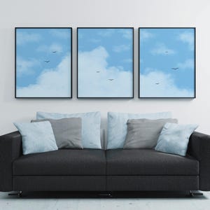 May include: Three framed art prints depicting a blue sky with white clouds and birds. They are hung above a dark grey sofa with light blue and grey cushions. The artwork provides a calming aesthetic to the room.