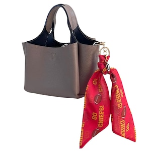 May include: A taupe leather handbag with a top handle and a detachable red scarf. The scarf is printed with footballs and the text "GO CHIEFS!". The bag has a structured design and a gold-tone metal clasp.