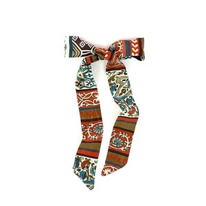 May include: A patterned hair tie with a bow in the center. The tie is made of a fabric with a brown, orange, blue, and white pattern.