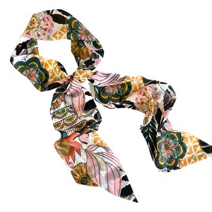 May include: A long, narrow scarf featuring a vibrant, multi-colored floral and geometric pattern. The design includes shades of deep green, mustard yellow, soft pink, and black, arranged in repeating mandala-like motifs and leaf shapes.