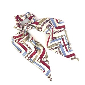 May include: A white scrunchie with a patterned scarf. The scarf features a repeating design of red, blue, and tan equestrian-style elements, including chains and straps. The scrunchie is a hair accessory.