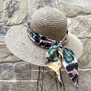 May include: A wide-brimmed straw hat with a woven texture, featuring a decorative scarf tied around the crown. The scarf displays a vibrant pattern in shades of dark green, mustard yellow, white, and black, with floral and geometric motifs.