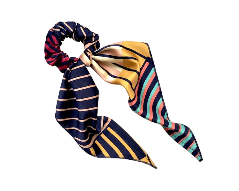 May include: A hair scrunchie with a long, flowing scarf tail. The scrunchie is dark red and black striped, while the scarf features a mix of navy, mustard yellow, coral pink, and teal stripes and geometric patterns. The scarf is tied in a bow around the scrunchie. This stylish hair accessory is perfect for adding a touch of elegance and flair to any outfit.