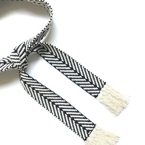 May include: Black and white woven belt with fringe ends. The belt is made of a thick, textured fabric and has a chevron pattern.
