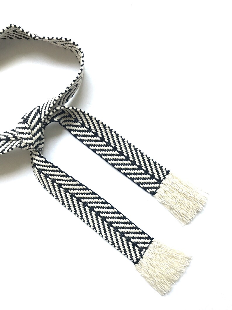 May include: Black and white woven belt with fringe ends. The belt is made of a thick, textured fabric and has a chevron pattern.