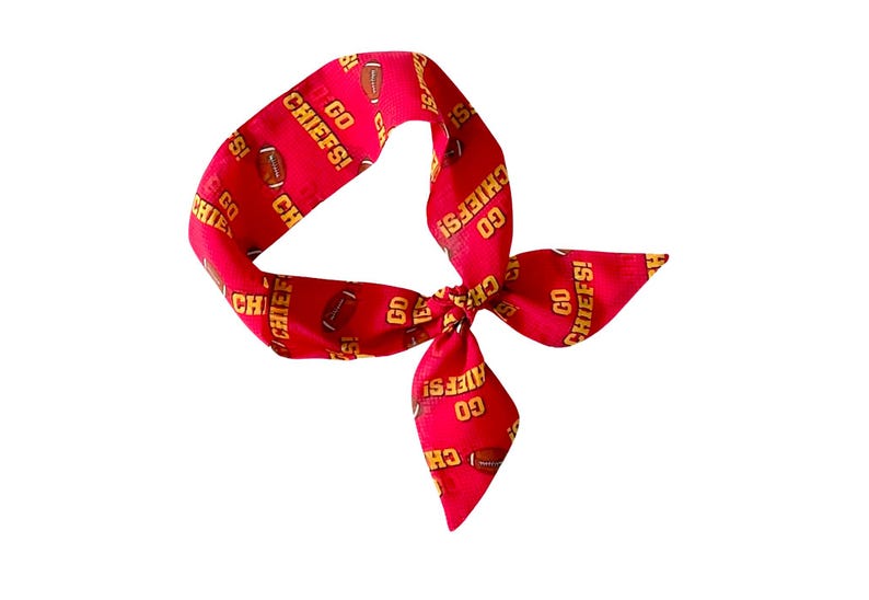 May include: A red fabric hair accessory featuring a repeating pattern of brown footballs and the yellow text GO CHIEFS!. The accessory is tied in a knot, creating a looped shape with two pointed ends extending from the center knot.