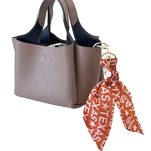 May include: Taupe-colored leather handbag with black interior and handles. A gold-toned keychain holds a scarf with the word "TEXAS" and star graphics in white and burnt orange.