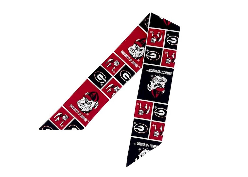 Puede incluir: Una cinta de tela larga y estrecha con un patr&oacute;n repetitivo de cuadrados rojos y negros. El dise&ntilde;o incluye el logotipo de la mascota bulldog de la University of Georgia y el texto University of Georgia impreso en blanco sobre fondos rojos y negros.