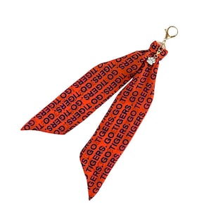 May include: Orange ribbon keychain with the text "GO TIGERS" in navy blue. The keychain has a gold-colored clasp and a small charm with a paw print. The ribbon is folded into two long, flowing sections.