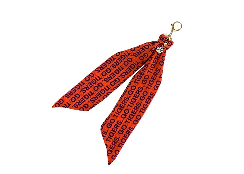 May include: Orange ribbon keychain with the text "GO TIGERS" in navy blue. The keychain has a gold-colored clasp and a small charm with a paw print. The ribbon is folded into two long, flowing sections.