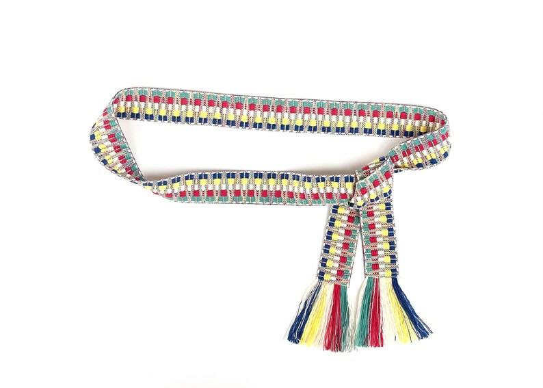 Puede incluir: Un cintur&oacute;n de tela tejida con un patr&oacute;n geom&eacute;trico repetitivo en hilos rojos, amarillos, azules y verdes sobre un fondo plateado. El cintur&oacute;n est&aacute; anudado y rematado con largos flecos multicolores en ambos extremos de la tira.