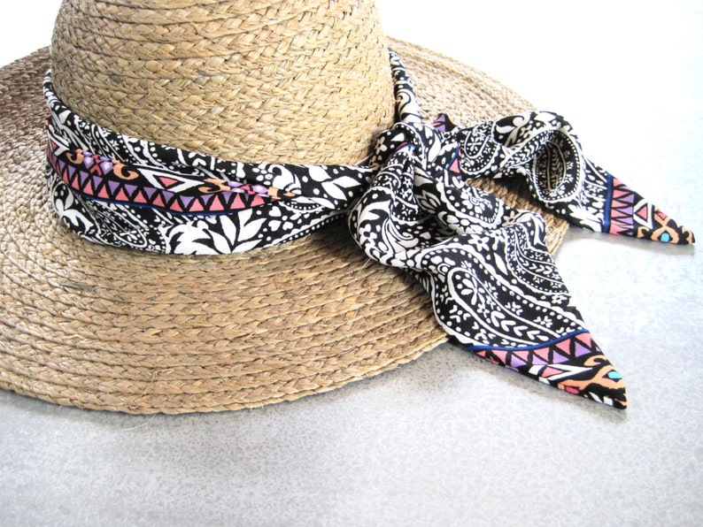 May include: A straw hat with a black and white paisley patterned ribbon tied around it. The ribbon has pink, blue, and orange accents.