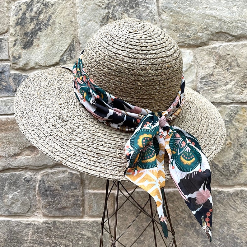 May include: A wide-brimmed straw hat with a woven texture, featuring a decorative scarf tied around the crown. The scarf displays a vibrant pattern in shades of dark green, mustard yellow, white, and black, with floral and geometric motifs.