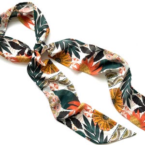 May include: A long, narrow hair scarf featuring a white background with a repeating pattern of dark green leaves, orange and yellow stylized flowers, and intricate black and white geometric details. The fabric has a smooth, slightly reflective finish.