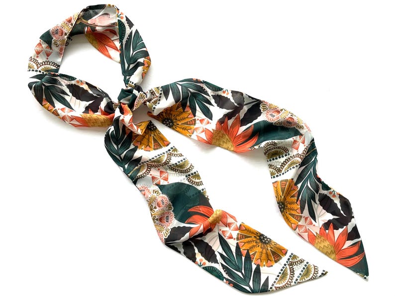 May include: A long, narrow hair scarf featuring a white background with a repeating pattern of dark green leaves, orange and yellow stylized flowers, and intricate black and white geometric details. The fabric has a smooth, slightly reflective finish.