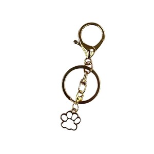 May include: Gold-colored keychain with a lobster clasp, a large ring, and a small charm shaped like a paw print. The paw print charm is white with a gold outline. The keychain is designed for attaching keys or other small items.