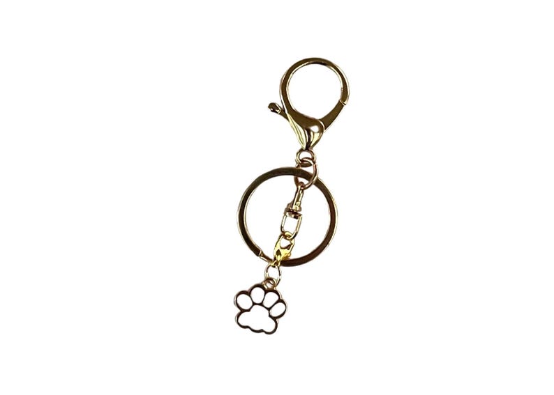 May include: Gold-colored keychain with a lobster clasp, a large ring, and a small charm shaped like a paw print. The paw print charm is white with a gold outline. The keychain is designed for attaching keys or other small items.