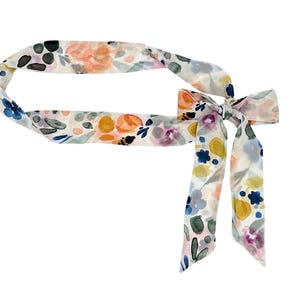 May include: A floral patterned hair scarf with a bow. The scarf features a watercolor design with orange, yellow, blue, and purple flowers on a white background. The scarf is tied in a bow.