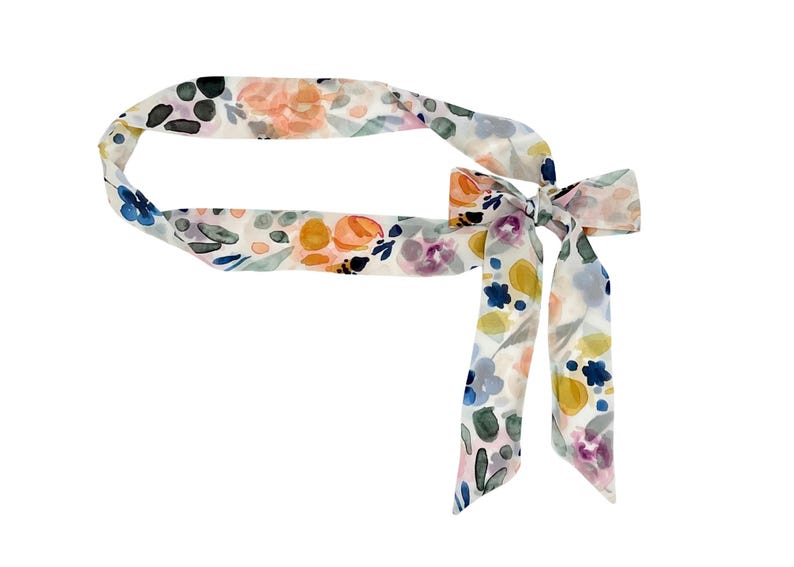 May include: A floral patterned hair scarf with a bow. The scarf features a watercolor design with orange, yellow, blue, and purple flowers on a white background. The scarf is tied in a bow.