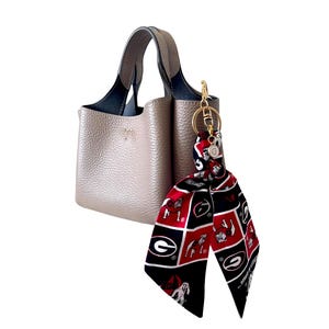 Puede incluir: Un bolso de mano color topo con superficie texturizada y dos asas. Sujeto a una asa hay un llavero de metal dorado con una cinta de tela decorativa con un patr&oacute;n de cuadr&iacute;cula rojo, negro y blanco que muestra una letra G y un bulldog.