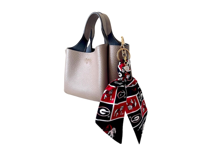 Puede incluir: Un bolso de mano color topo con superficie texturizada y dos asas. Sujeto a una asa hay un llavero de metal dorado con una cinta de tela decorativa con un patr&oacute;n de cuadr&iacute;cula rojo, negro y blanco que muestra una letra G y un bulldog.