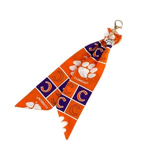 May include: Orange and purple Clemson Tigers keychain accessory. The fabric features the letter "C" in purple squares, white paw prints, and the word "CLEMSON" in white. A gold-colored clasp is attached.