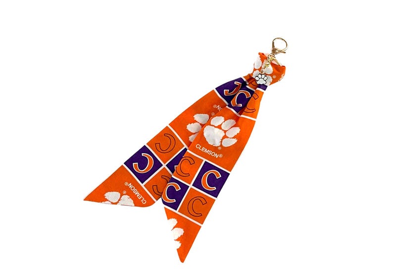 May include: Orange and purple Clemson Tigers keychain accessory. The fabric features the letter "C" in purple squares, white paw prints, and the word "CLEMSON" in white. A gold-colored clasp is attached.