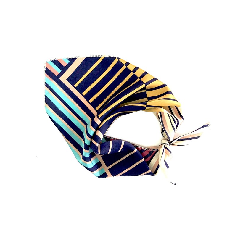 May include: A navy blue, yellow, pink, and turquoise patterned bandana with a geometric design. The bandana is tied in a knot.