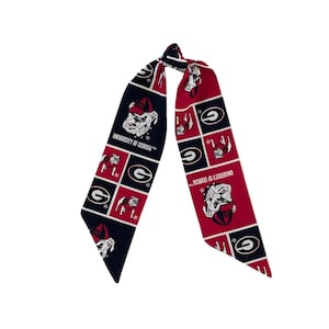 Puede incluir: Una bufanda larga y estrecha con un patr&oacute;n de cuadr&iacute;cula repetitivo en rojo, negro y blanco. El dise&ntilde;o incluye el texto University of Georgia, la mascota bulldog con gorra roja y el logotipo de la letra G impreso en la tela.